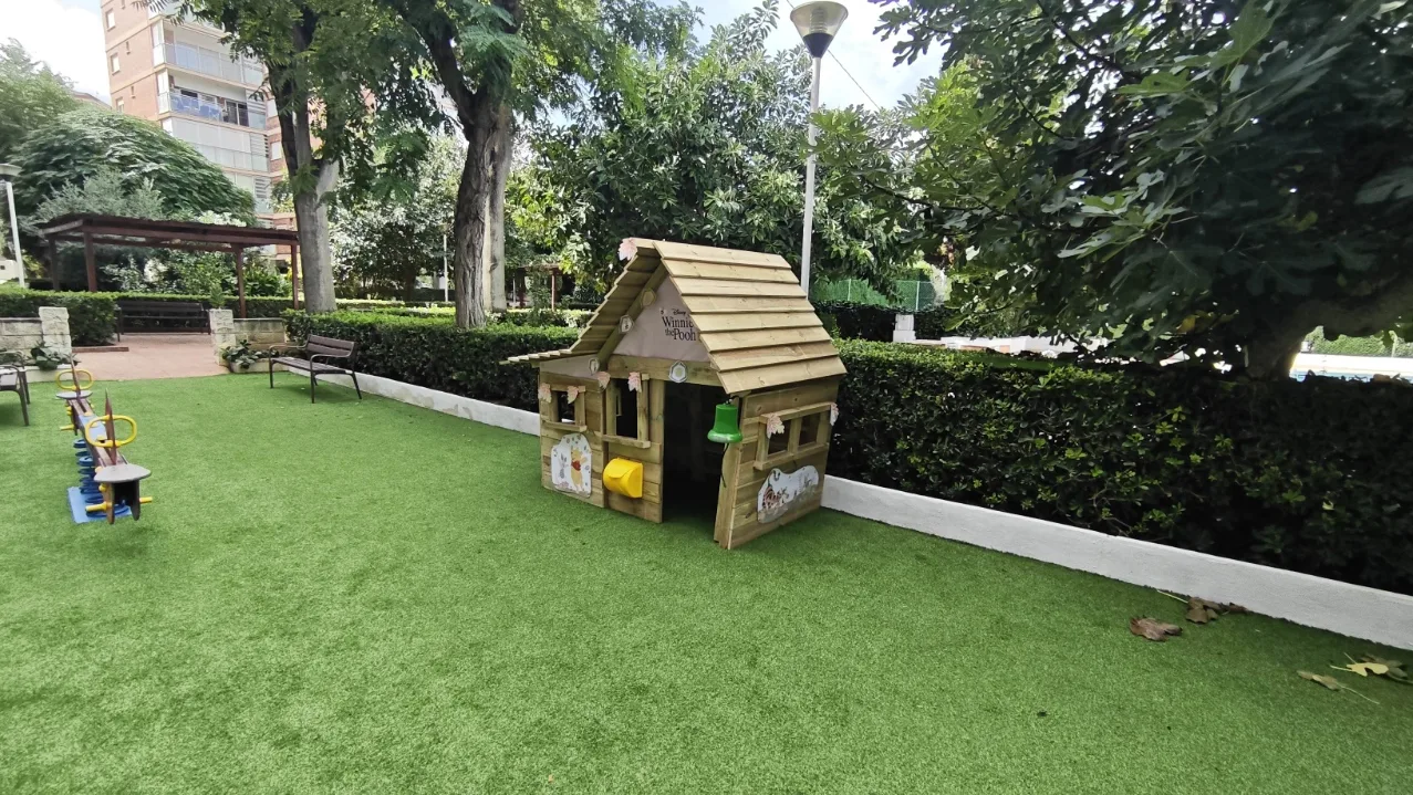 Casita, balancín y bancos sobre césped artificial en un parque infantil comunitario de Dénia