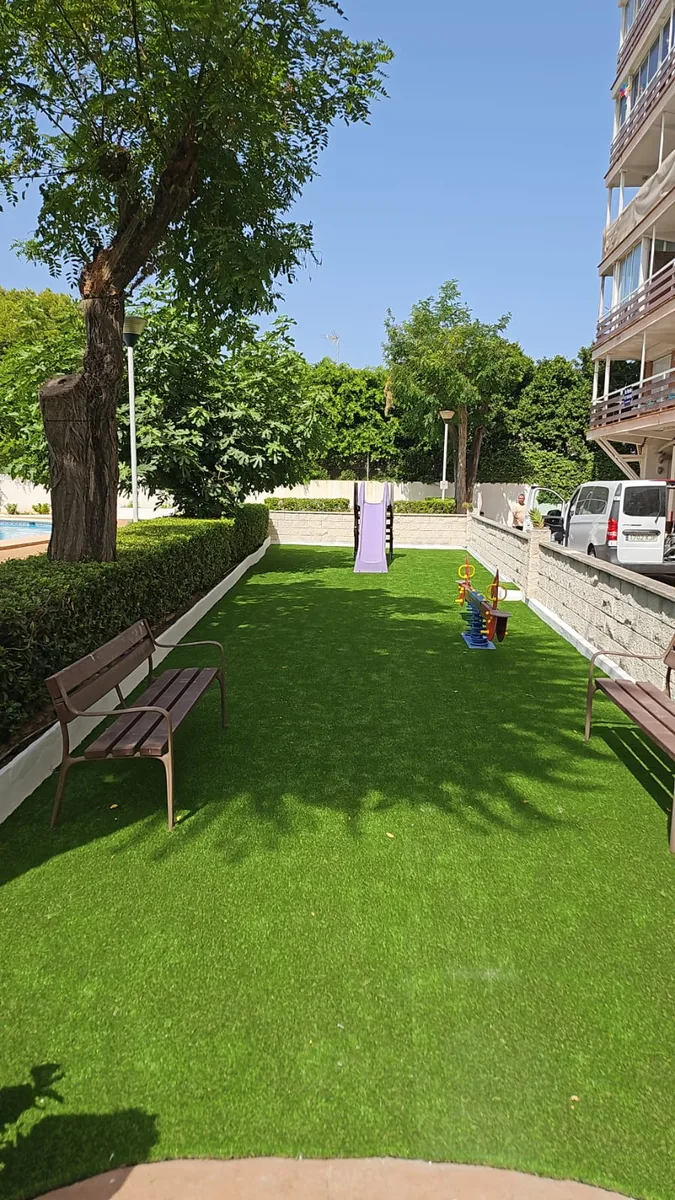Vista general del parque infantil de Dénia con césped artificial, bancos y tobogán