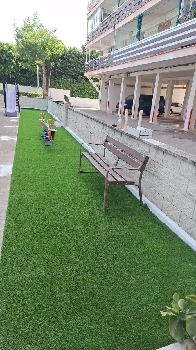 Césped artificial AMORE 22 mm junto al muro y el banco de un parque infantil de Dénia